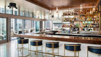 A modern bar with high stools, a wooden counter, and shelves stocked with bottles; large windows offer a city view.