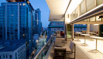 A modern cityscape view from a balcony with elegant seating, overlooking tall buildings and a street illuminated by evening lights.