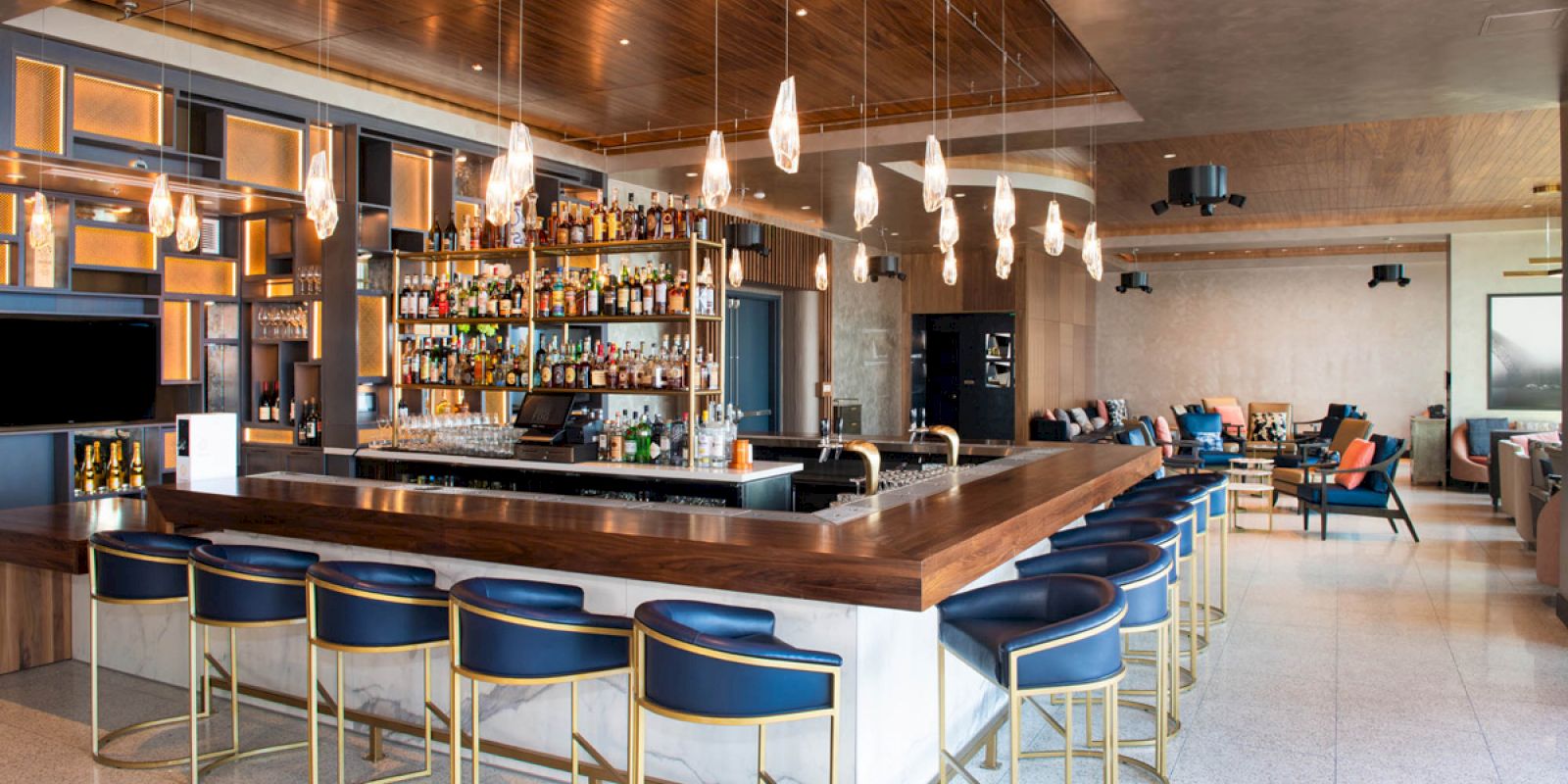 Modern bar interior with wooden accents, hanging lights, a fully stocked bar, and blue cushioned stools. Cozy seating area in the background.