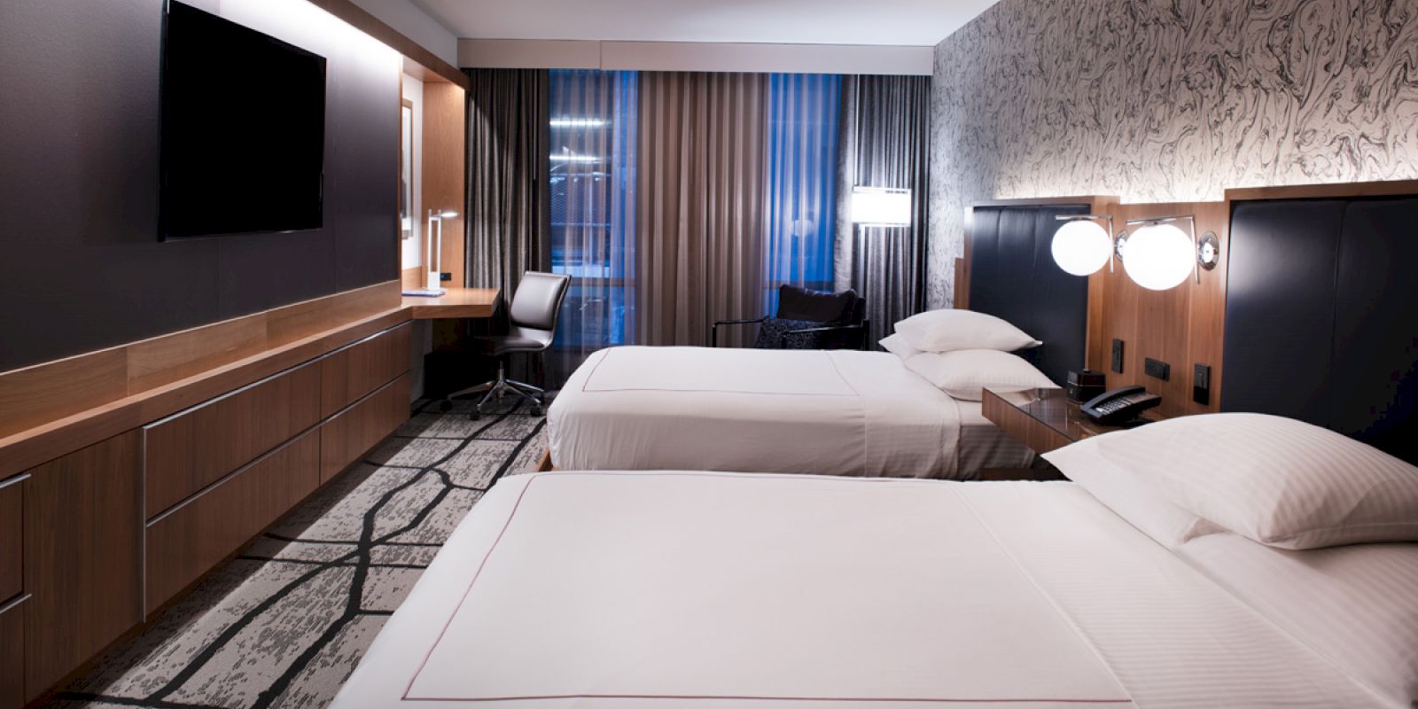 This image shows a modern hotel room with two beds, a TV, desk, chair, and stylish decor, featuring patterned wallpaper and curtains.