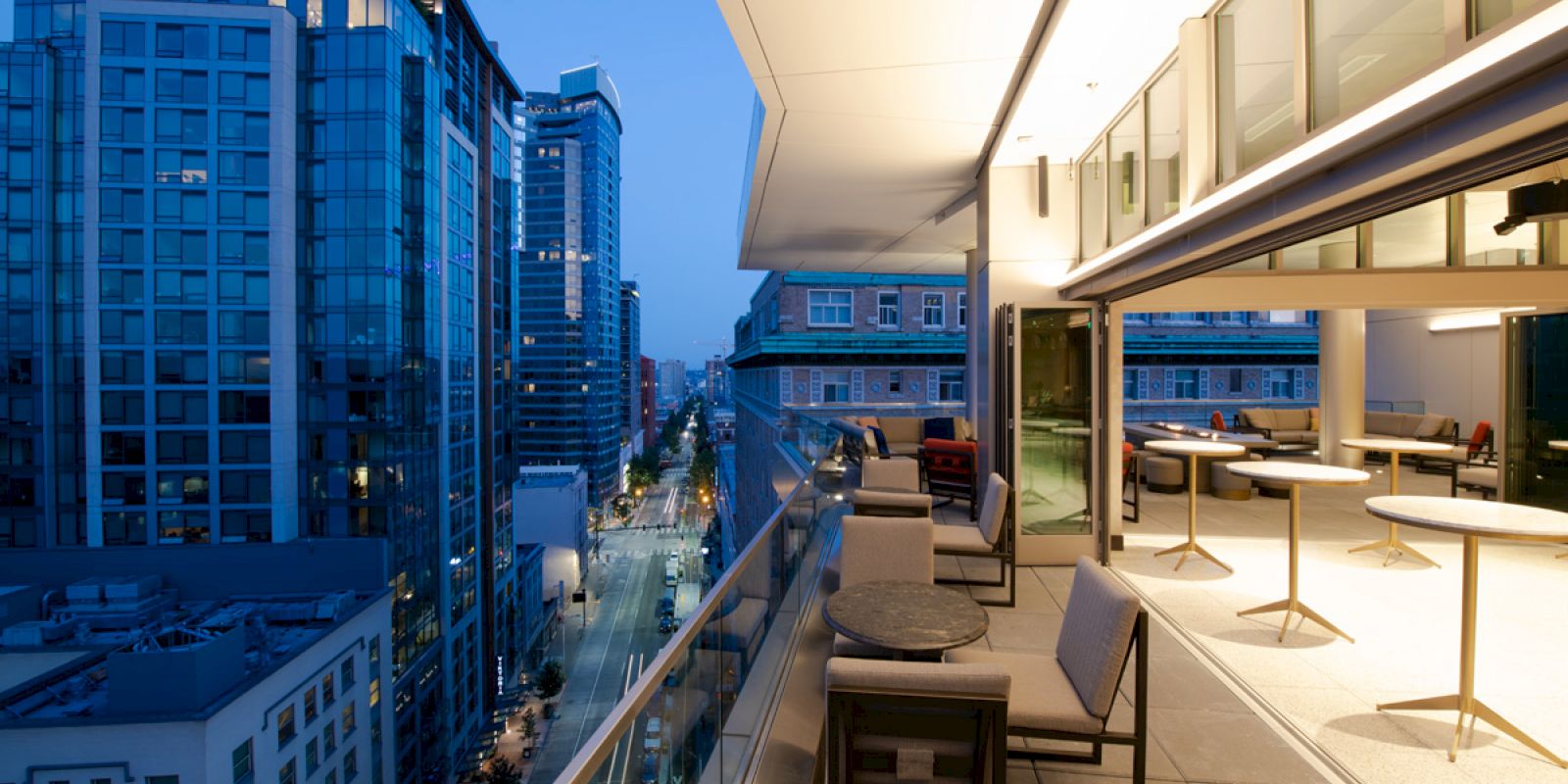 City view from a modern balcony at dusk, showing a row of tall buildings and a partially visible interior with tables and chairs.