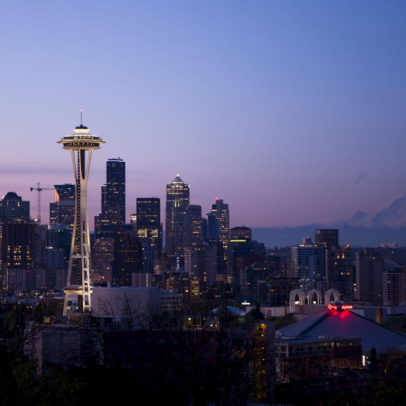 Seattle skyline at dusk featuring the Space Needle; Mount Rainier visible in the background under a purple and blue sky.