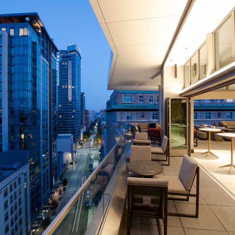 A modern cityscape with tall buildings is seen from a sleek outdoor terrace featuring tables and chairs, with city lights glowing below.
