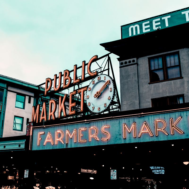 The image shows a public market with neon signs for a farmers market, featuring a large clock, and buildings in the background.