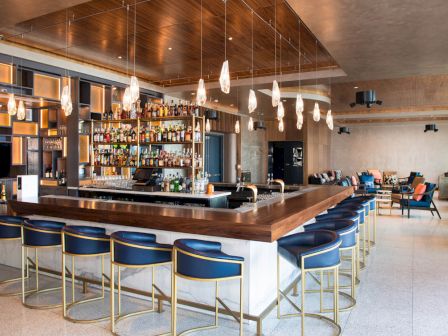 A modern bar with a sleek countertop, blue stools, hanging lights, and a well-stocked liquor shelf, in a spacious lounge area.