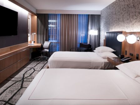 The image shows a modern hotel room with two beds, a wall-mounted TV, a work desk, and stylish decor.