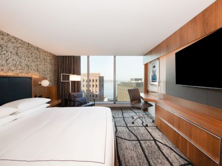 A modern hotel room with a large bed, desk, chair, big TV, and a window view of buildings and water.
