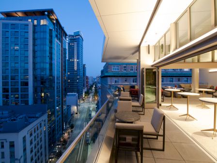 A modern rooftop terrace with outdoor seating overlooks a cityscape featuring tall buildings and a street below, captured at dusk.