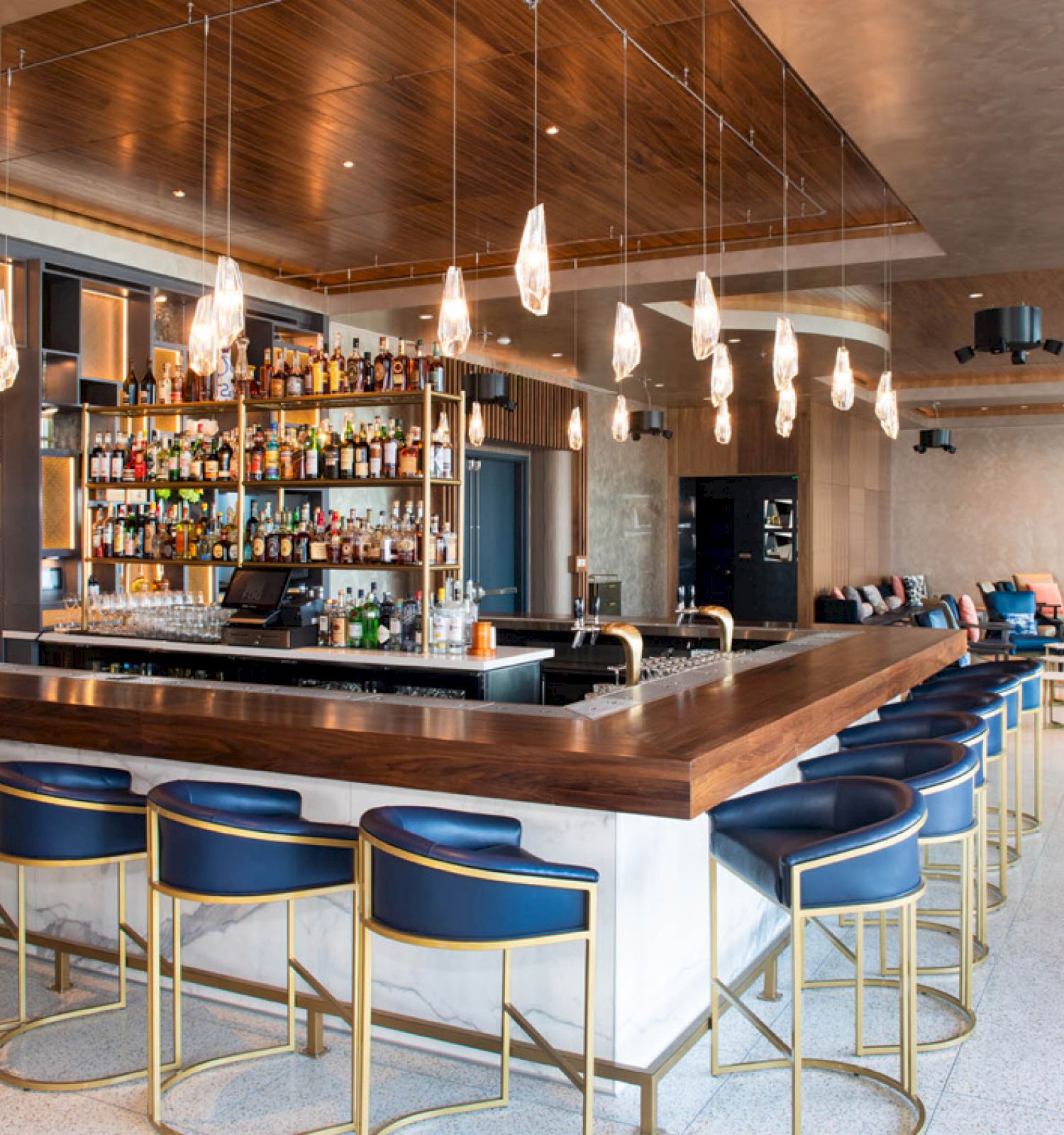 A modern bar with blue stools, a variety of bottles behind the counter, and stylish lighting in a spacious, contemporary setting.