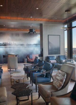 A modern lounge with plush seating, cityscape wall art, and large windows offering a view of a waterfront and Ferris wheel during daytime.