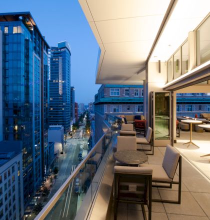 A modern cityscape at dusk with tall buildings and a stylish rooftop terrace featuring seating and tables.