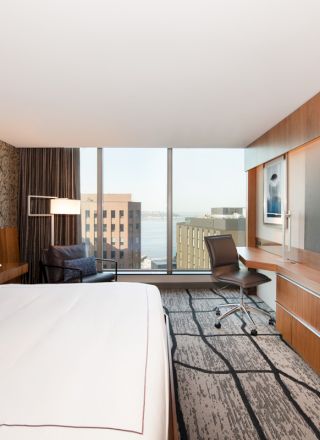 A modern hotel room with a bed, wall-mounted TV, desk, chair, and large window with a city view, featuring contemporary decor.