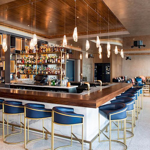 A modern bar with blue stools, hanging lights, and a stocked shelf sits in a stylish, open lounge area with additional seating.