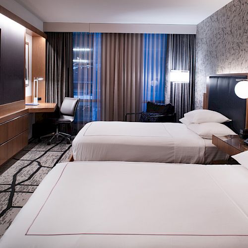 The image shows a modern hotel room with two beds, a large TV, a desk with a chair, and stylish decor.