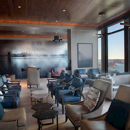 A modern lounge with numerous chairs, a mural on the wall, and large windows offering a scenic waterfront and city view at sunset.