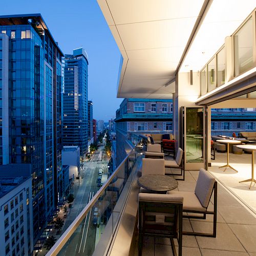 A modern rooftop terrace with seating overlooks a cityscape featuring tall buildings and illuminated streets at twilight.