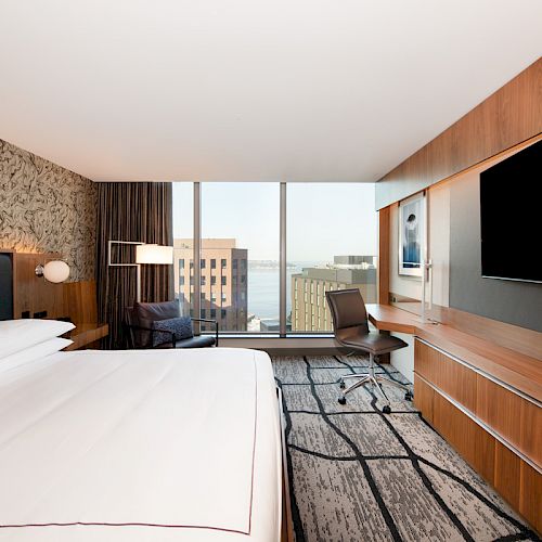 A modern hotel room with a large bed, wall-mounted TV, desk, and city view through a window, featuring contemporary decor.
