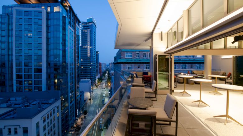 A modern rooftop terrace with seating overlooks a cityscape at dusk, featuring tall buildings and illuminated streets in the background.