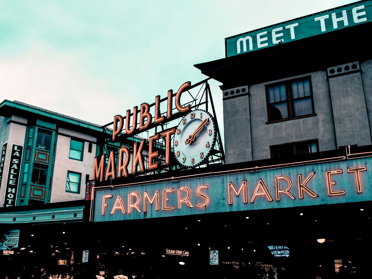 The image shows a classic neon sign for a "Public Market" and "Farmers Market" on a building with a clock, under a cloudy sky.