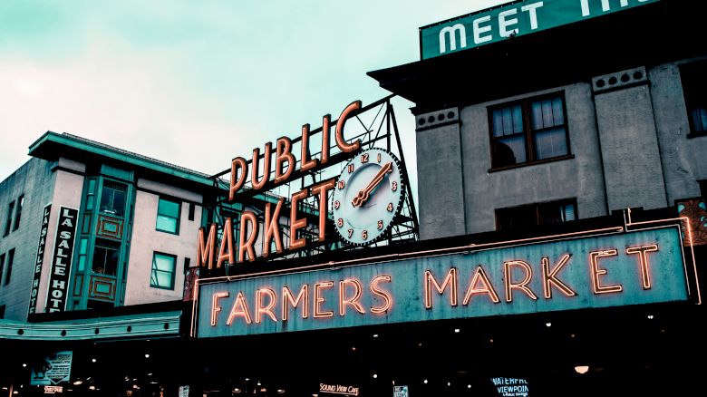 The image shows a classic neon sign for a "Public Market" and "Farmers Market" on a building with a clock, under a cloudy sky.