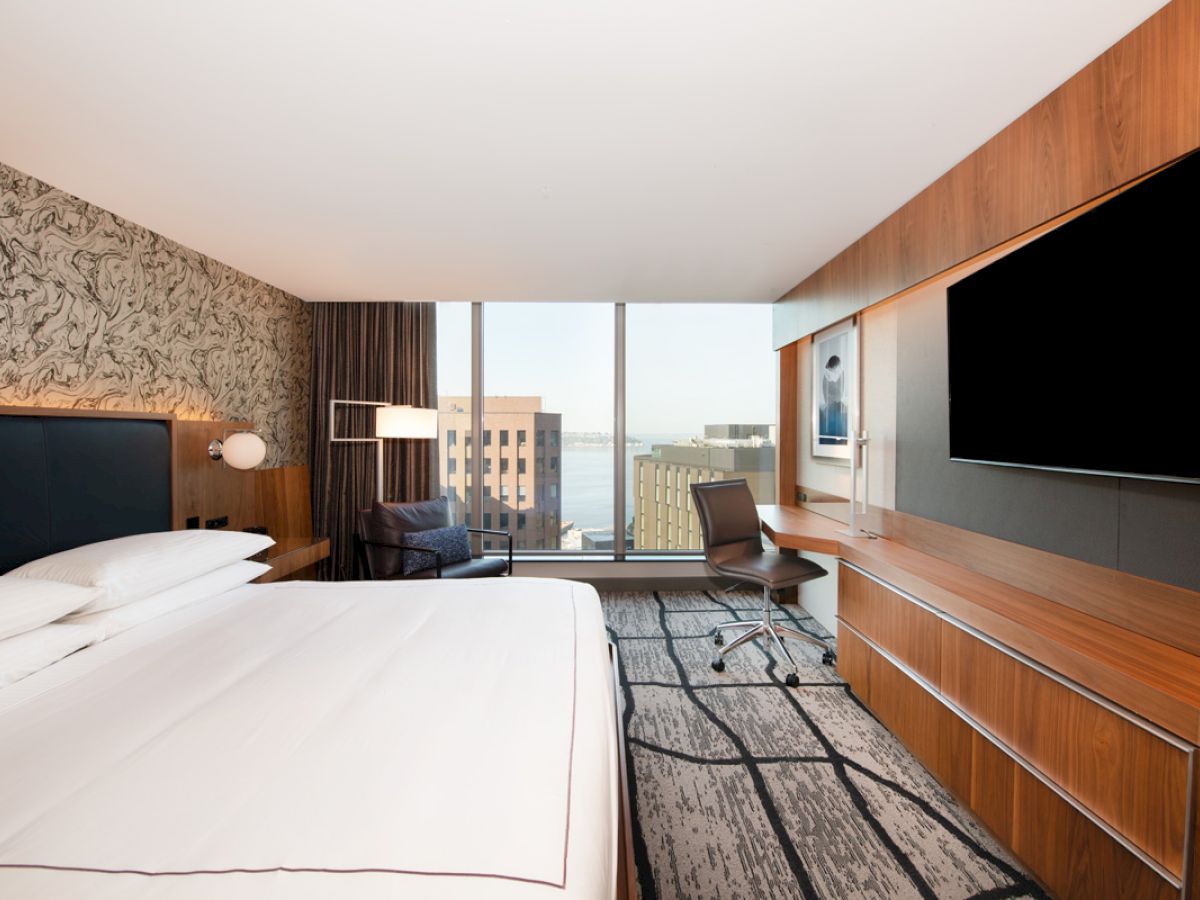 A modern hotel room with a large bed, desk, chair, and TV. The window offers a view of buildings and water.