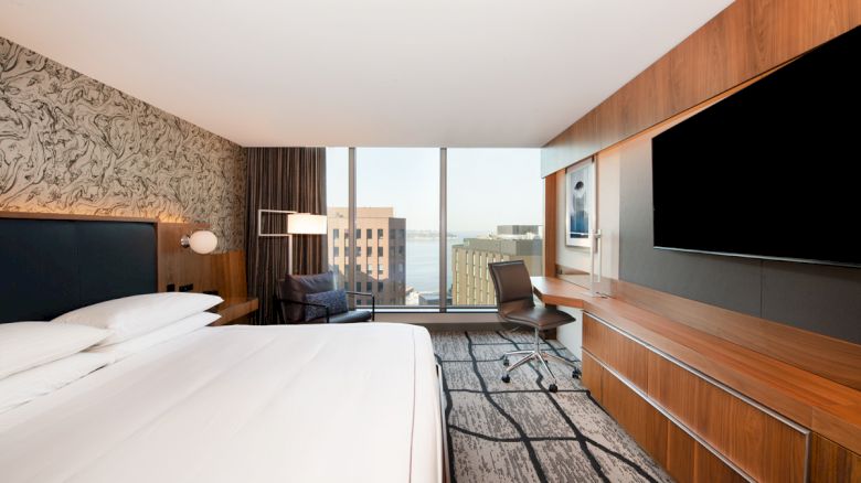 A modern hotel room with a large bed, desk, chair, and TV. The window offers a view of buildings and water.