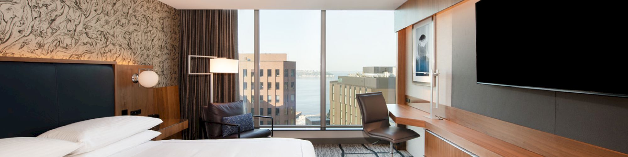 A modern hotel room with a large bed, TV, desk, and chair, offering a city view through floor-to-ceiling windows.