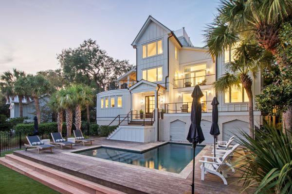 A modern house with large windows, surrounded by trees, features a pool and seating area with loungers and umbrellas.