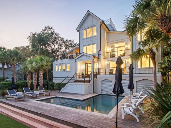 A modern house with large windows, surrounded by trees, features a pool and seating area with loungers and umbrellas.